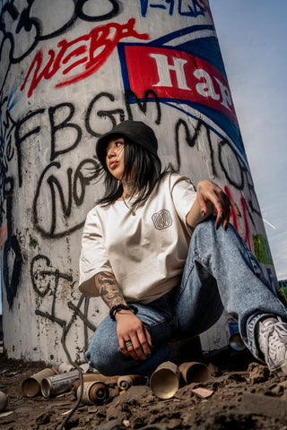 Model wearing the off white vindicated tee shirt with vindicated logo on chest.  Model is kneeling wearing denim jeans and black bucket hat with white shoes.