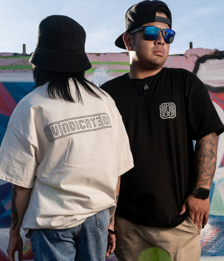 Two models, one facing backwards wearing the off white vindicated tee shirt with Vindicated written on back and one model facing forward wearing black Vindicated tee shirt with logo on the front.